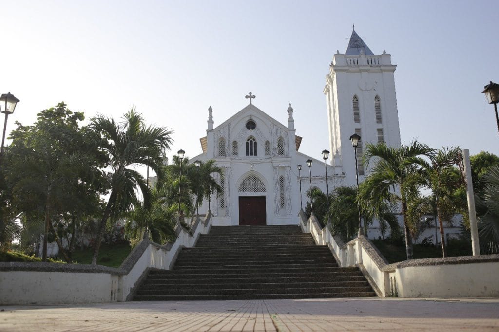 Iglesia de fachada blanca con torre campanario, escalinatas y palmeras en un entorno urbano del departamento de Bolívar.