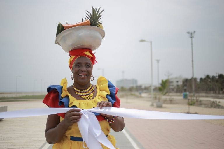 Mujer con vestimenta tradicional afrocolombiana sosteniendo una cinta ceremonial y llevando una batea con frutas sobre la cabeza durante un acto cultural.