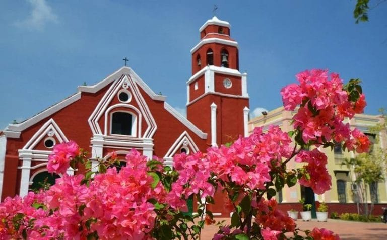 Iglesia San Francisco en Santa Cruz de Mompox, Bolívar, con fachada roja y flores rosadas en primer plano.